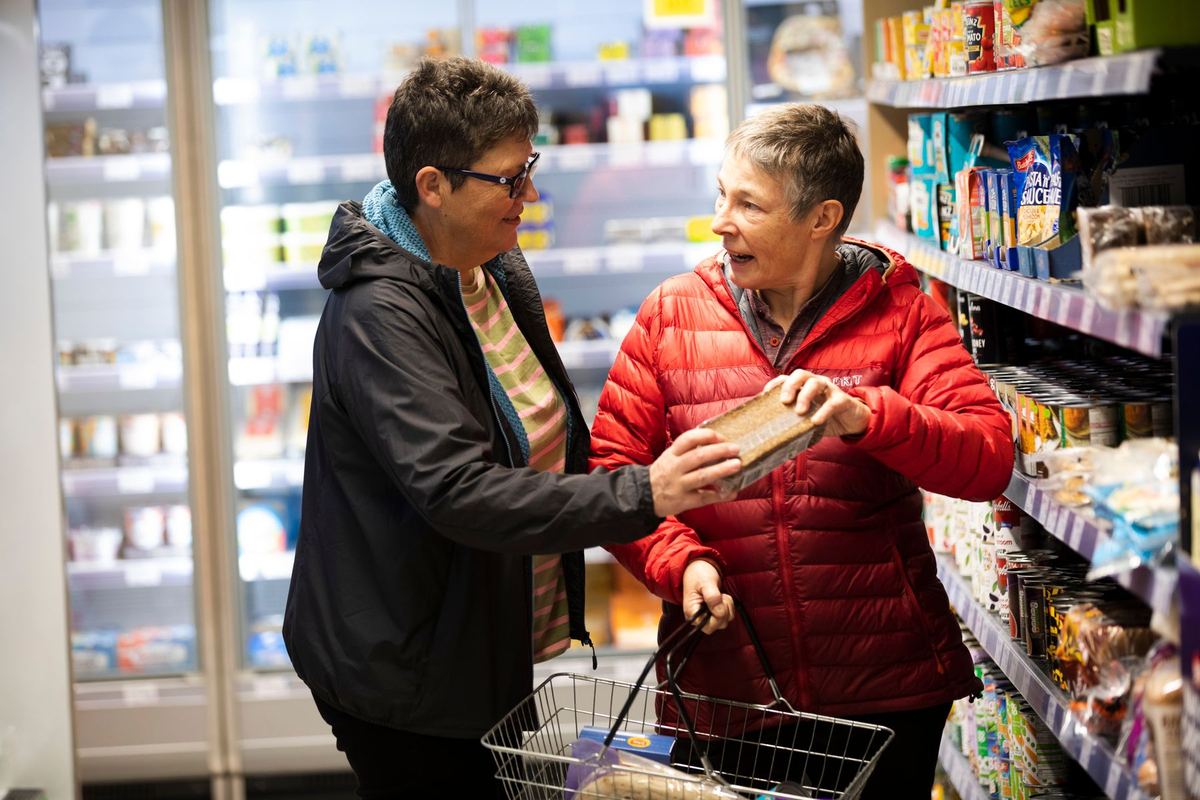 Two older women talking over a product in a supermarket aisle